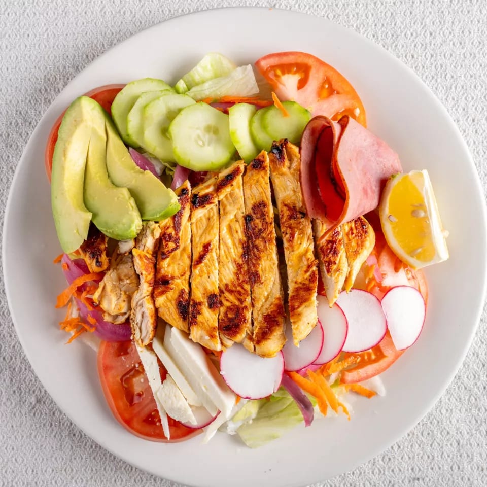 Grilled chicken salad with avocado, cucumber, tomato, ham, radish, and lemon on a white plate.