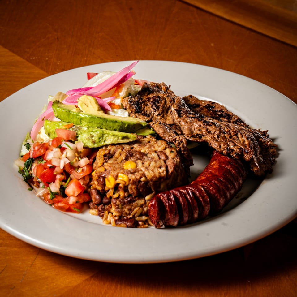 Grilled steak, sausage, rice and beans, pico de gallo, avocado, and pickled onions on a white plate.