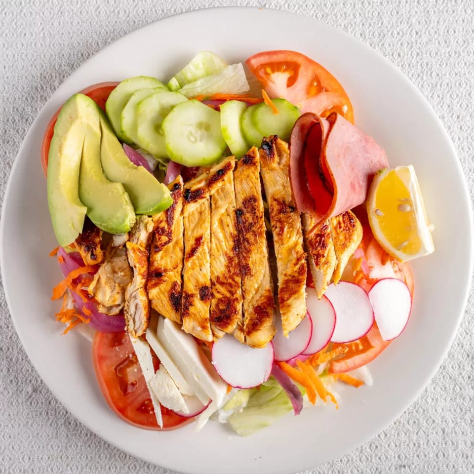 Grilled chicken salad with avocado, cucumber, tomato, radish, ham, and a lemon wedge.