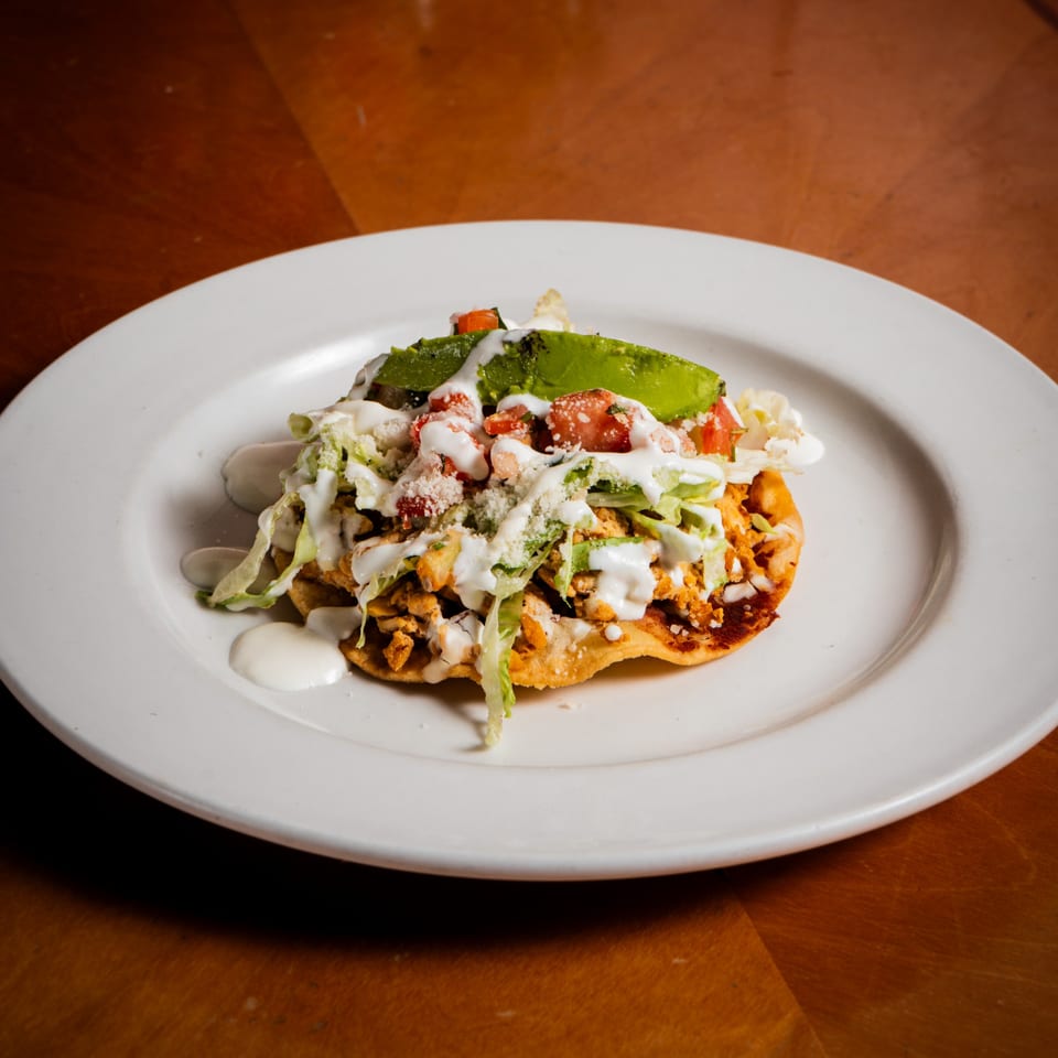 A chicken tostada with lettuce, tomato, avocado, cheese, and sour cream on a white plate.