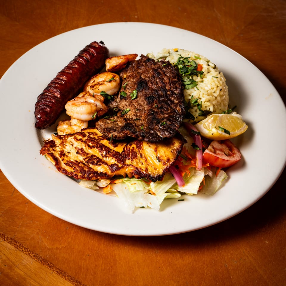 Grilled sausage, steak, shrimp, and chicken with rice, salad, and lemon on a white plate.